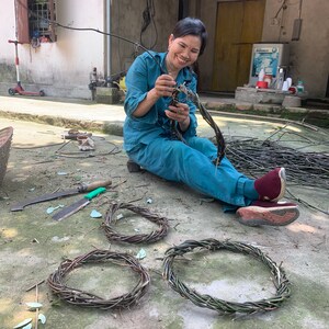 Puede incluir: Una mujer se sienta en un patio de concreto y teje ramas en coronas circulares. Tres coronas est&aacute;n dispuestas en el suelo.