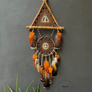 May include: A brown and orange dreamcatcher with feathers hanging from the bottom. The dreamcatcher is made with a wooden triangle frame and a circular hoop. The dreamcatcher is hanging from a string against a gray wall.