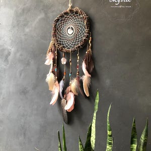 Puede incluir: Un atrapasue&ntilde;os marr&oacute;n con plumas y cuentas que cuelgan de &eacute;l. El atrapasue&ntilde;os est&aacute; hecho de materiales naturales y tiene un dise&ntilde;o tejido blanco y marr&oacute;n. El texto "Mystic Handmade by Kham" es visible en la esquina superior derecha de la imagen.