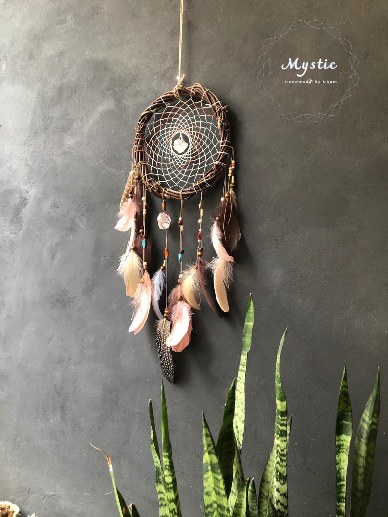 Puede incluir: Un atrapasue&ntilde;os marr&oacute;n con plumas y cuentas que cuelgan de &eacute;l. El atrapasue&ntilde;os est&aacute; hecho de materiales naturales y tiene un dise&ntilde;o tejido blanco y marr&oacute;n. El texto "Mystic Handmade by Kham" es visible en la esquina superior derecha de la imagen.