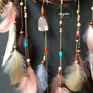 Puede incluir: Un atrapasue&ntilde;os con ramas de madera marr&oacute;n, un dise&ntilde;o de plumas rosa, blanco y marr&oacute;n, y un cristal rosa colgando del centro. El atrapasue&ntilde;os tiene una variedad de cuentas en marr&oacute;n, blanco, rojo y azul.