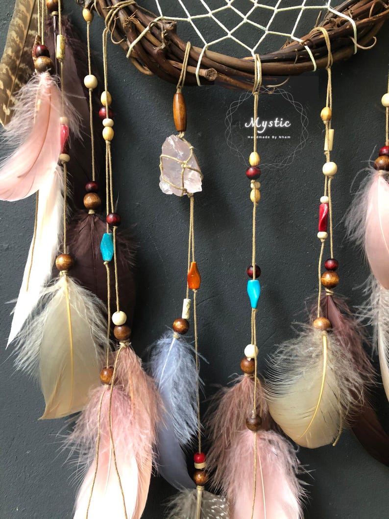 Puede incluir: Un atrapasue&ntilde;os con ramas de madera marr&oacute;n, un dise&ntilde;o de plumas rosa, blanco y marr&oacute;n, y un cristal rosa colgando del centro. El atrapasue&ntilde;os tiene una variedad de cuentas en marr&oacute;n, blanco, rojo y azul.