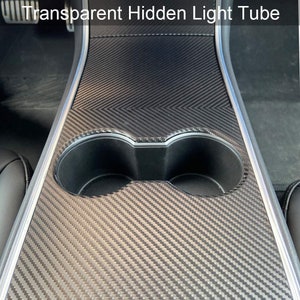 May include: A black carbon fiber console insert for a Tesla Model 3 or Y with two cup holders. The insert has a textured surface and a silver trim.