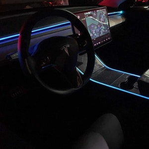 May include: The interior of a black Tesla car with a blue LED light strip running along the dashboard and center console. The car's steering wheel has a Tesla logo and the touchscreen display shows a map.
