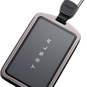 May include: A black metal key fob with a silver trim and a black leather strap. The fob has the word "TESLA" printed on it in silver lettering.