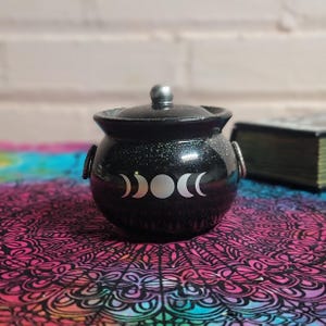 May include: A black, glitter-covered cauldron with a lid and silver handle. The cauldron features a white crescent moon phase design. The cauldron is sitting on a colorful, patterned surface.