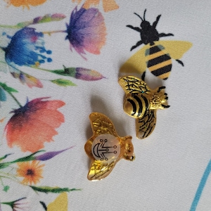 May include: Two gold bee-shaped charms with black and yellow stripes. One charm has a white center with a geometric design.
