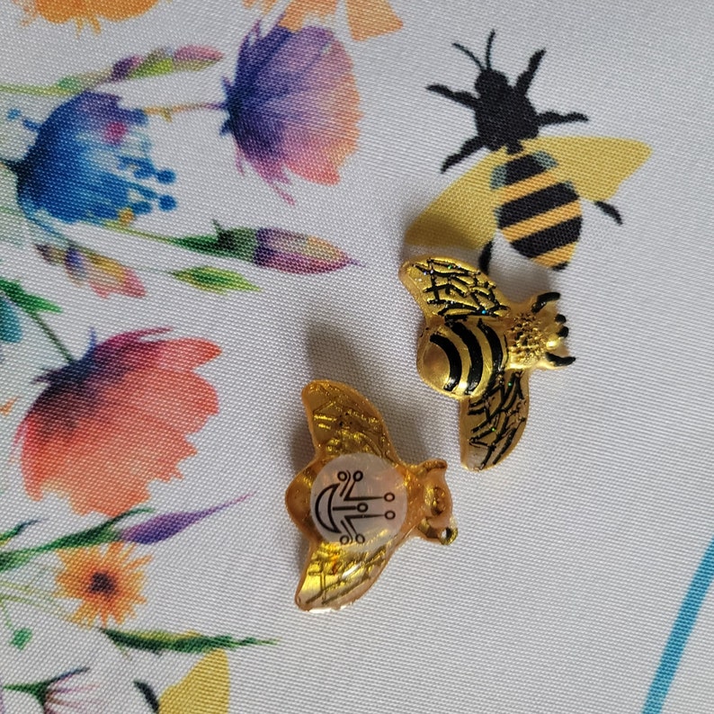 May include: Two gold bee-shaped charms with black and yellow stripes. One charm has a white center with a geometric design.