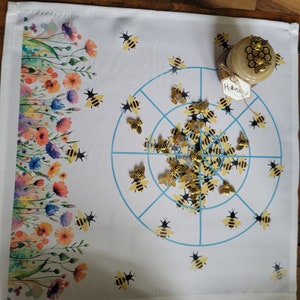 May include: A white fabric game board with a floral border and a blue and white target in the center.  The target has a series of concentric circles and lines.  There are small gold bee figurines scattered around the target.  A small glass jar with a gold lid and the word "Honey" printed on a label is in the upper right corner of the image.