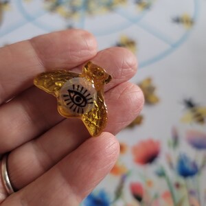 May include: A gold resin bee charm with a black eye design. The bee is translucent and has a glittery finish.
