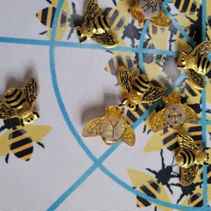 May include: A set of gold bee-shaped charms with different symbols on their backs. The charms are on a white background with a blue and white pattern.