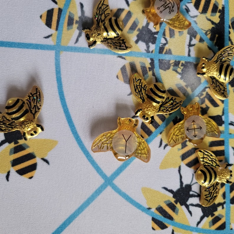 May include: A set of gold bee-shaped charms with different symbols on their backs. The charms are on a white background with a blue and white pattern.