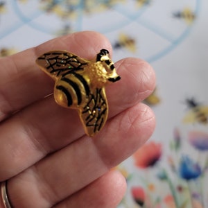 May include: A gold and black bee pin with glitter accents. The bee is in flight with its wings spread out.