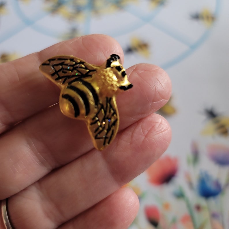 May include: A gold and black bee pin with glitter accents. The bee is in flight with its wings spread out.