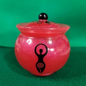 May include: A small, round, red jar with a lid and a black knob. The jar has a black silhouette of a goddess figure on the front. The jar is on a green surface.