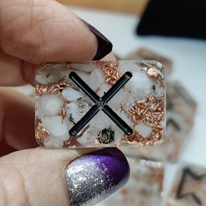 Peut inclure: Une petite tuile carrée en résine transparente avec un motif en X noir intégré. La tuile est remplie de petites paillettes blanches et dorées.