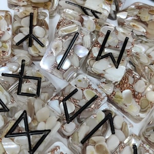 May include: A set of white rectangular tiles with black runes and iridescent shell pieces embedded in the resin. The tiles are scattered on a white surface.