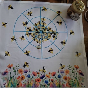 May include: A white fabric target with a blue circle and a grid pattern.  The target is decorated with black and yellow bees.  The text "Honey" is printed on a small tag in the upper right corner of the image.  A border of colorful flowers is at the bottom of the target.