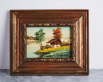 May include: A framed oil painting depicting a landscape scene with a small house, trees, and a lake. The painting is in a brown wooden frame with gold accents.