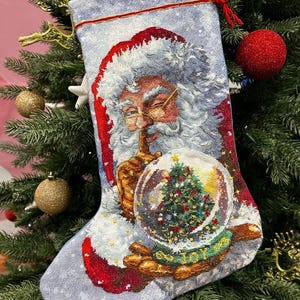 May include: Christmas stocking featuring Santa Claus holding a snow globe with a Christmas tree inside. The stocking has a light blue background with a red trim and a red loop for hanging. The image is set against a Christmas tree.