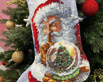 May include: Christmas stocking depicting Santa Claus holding a snow globe with a Christmas tree. The stocking has a light blue background, red trim, and a red hanging loop. The image is set against a Christmas tree.