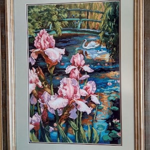 May include: Framed cross-stitch artwork depicting a serene water garden scene. Pink irises bloom in the foreground, with a bridge and swans on the water in the background. The artwork is framed in a gold-toned wooden frame.