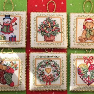 Może przedstawiać: Sześć kwadratowych ozdób świątecznych z haftem krzyżykowym. Każda ozdoba przedstawia inny motyw świąteczny, w tym bałwana, choinkę, misia, pończochy, wieniec i serce. Ozdoby mają złote sznurkowe wieszaki.