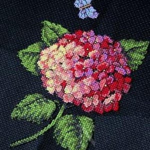 May include: Cross-stitch embroidery featuring a hydrangea flower with red, pink, and yellow petals, and green leaves. A small butterfly with blue and purple details is above the flower. The design is on a dark blue fabric.