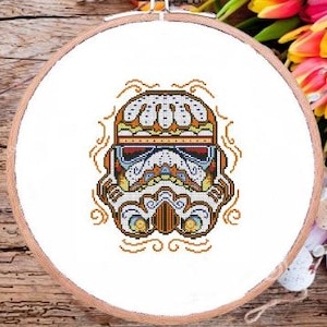 Peut inclure: Broderie au point de croix d'un casque de Stormtrooper dans un design de crâne de sucre du Jour des Morts. Le casque présente des motifs complexes en orange, jaune, bleu et blanc, sur fond blanc dans un cercle à broder en bois.