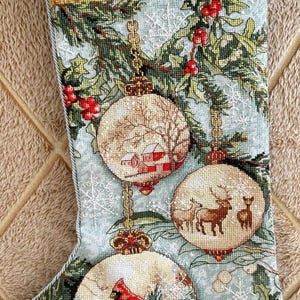 May include: Embroidered Christmas stocking with a pale blue backdrop and festive ornaments. The decorations depict winter scenes with a red cardinal, deer, and a snowy village. Holly and snowflakes adorn the stocking.