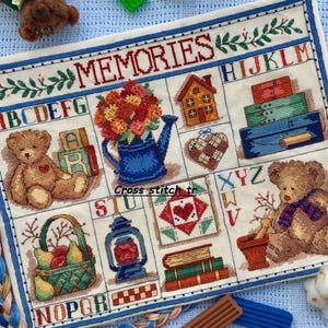 May include: Cross-stitch sampler with the word "MEMORIES" and various embroidered images, including teddy bears, flowers, a house, and a basket of fruit. The sampler has a blue border and is on a light blue background.