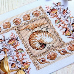 Könnte beinhalten: Kreuzstich-Stickerei mit nautischem Thema, die Muscheln, Fische und Korallen darstellt. Das zentrale Design zeigt eine Nautilus-Schale und das Wort "Nautilus". Das Kunstwerk ist mit einem dekorativen Rahmen und dem Text "Cross Stitch TR" versehen.