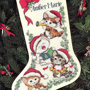 May include: A Christmas stocking with embroidered kittens wearing Santa hats, playing musical instruments, and surrounded by a green wreath. The name "Amber Marie" is embroidered in blue. A red bow and ribbon are at the top.