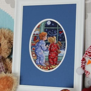 May include: A framed cross-stitch artwork featuring two children in pajamas gazing out a window at a starry night scene. The artwork is set against a blue background within a white frame. The children are in blue and red pajamas.