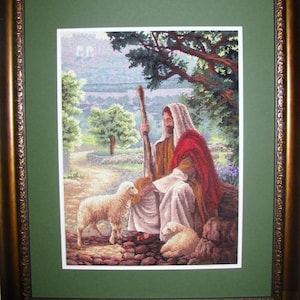 May include: Framed cross-stitch artwork showing a figure in white and red robes with a staff, seated near two lambs. The pastoral scene includes a path, trees, and a distant building, within a gold-toned frame.