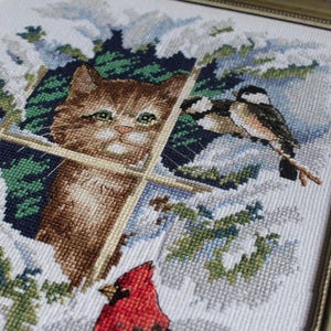 Puede incluir: Obra de arte de punto de cruz en un marco plateado. La imagen representa un gato atigrado marrón mirando por una ventana a pájaros en una escena nevada. Un cardenal rojo está posado debajo. La obra de arte es detallada y colorida.