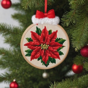 Puede incluir: Un adorno de Navidad con forma de bola con una poinsettia roja bordada en una tela blanca. La bola está adornada con un borde de piel sintética blanca y un lazo rojo.