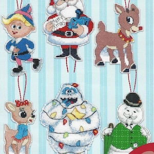 May include: A collection of Christmas ornaments featuring cross-stitch designs. The ornaments include Santa Claus holding a toy train, a reindeer, a snowman, and an elf. The Santa ornament has the text "Merry Xmas". The ornaments are on a blue and white striped background.