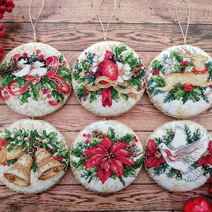 Puede incluir: Seis adornos navideños redondos con diseños bordados. Cada adorno presenta un tema festivo diferente, incluyendo pájaros, un cardenal, un reno, campanas, una flor de pascua y una paloma. Los adornos tienen un fondo color crema y están adornados con una cuerda dorada para colgar.