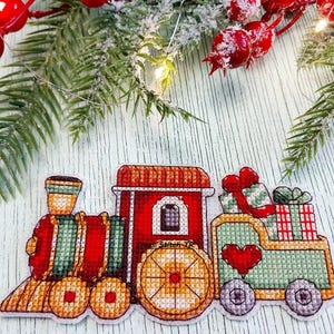 Könnte beinhalten: Ein Kreuzstich-Weihnachtszug mit roten, grünen und goldenen Akzenten. Der Zug hat eine rote Lokomotive, einen roten Personenwagen und einen grünen Wagen mit Geschenken. Ein rotes Herz befindet sich auf dem grünen Wagen. Festliches Urlaubsthema.