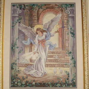 Op de afbeelding: Kruissteekkunstwerk van een engel met uitgespreide vleugels, die een kaars vasthoudt. De engel draagt een witte jurk en een blauwe sjerp, voor een stenen boog. Het kunstwerk is ingelijst in een goudkleurige lijst.