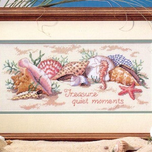 Op de afbeelding: Kruissteekkunstwerk in een houten lijst. De afbeelding toont verschillende schelpen, een zeepaardje en een zeester op een zanderige achtergrond. De tekst "Treasure quiet moments" is onder de zeedieren geborduurd. De lijst is bruin.