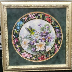 May include: A framed needlepoint artwork. The central circular design depicts a hummingbird amidst purple, white, and pink flowers. The artwork is framed in a gold-coloured, ornate frame, set against a dark green background. The artwork is a decorative piece.