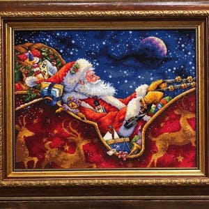 May include: A framed cross-stitch embroidery depicting Santa Claus in a red suit with a white beard, driving a sleigh filled with toys against a dark blue starry night sky with a large moon. The sleigh features gold reindeer silhouettes.