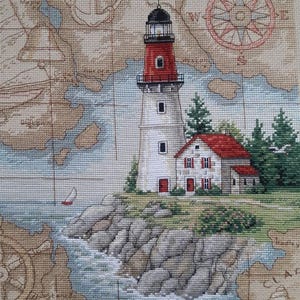 May include: Cross-stitch of a lighthouse with a red top and a small house with a red roof, set on a rocky coastline. The background features a vintage map with a compass rose and an anchor.
