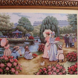 May include: A framed cross-stitch depicting a park scene with figures in period clothing. The artwork features a pond, a carousel, and lush greenery, framed by a decorative gold border. The scene is filled with people enjoying a sunny day.