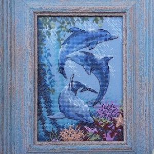May include: Cross-stitch artwork of three blue dolphins swimming in the ocean, framed in a distressed blue wooden frame. The underwater scene includes coral and plants in shades of purple, orange, and green.
