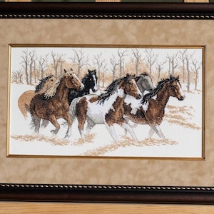May include: Framed cross-stitch artwork showing a herd of horses running through a snowy landscape. The horses are various shades of brown, black, and white. The artwork is framed in a dark brown wooden frame with a light brown mount.