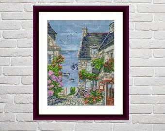 May include: A framed cross-stitch embroidery showing a charming seaside village with colourful houses, lush greenery, and a view of the sea. The scene includes a bicycle parked on a cobbled street.