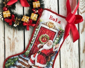 May include: A festive Christmas stocking with the name "Buka" embroidered on it, featuring a detailed image of Santa Claus and a red door. A Christmas wreath and red ribbon are also visible.
